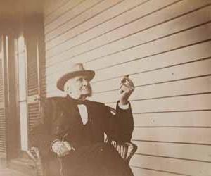 西德尼·巴特利特坐在门廊的椅子上，面朝左，拿着一张雪茄照片